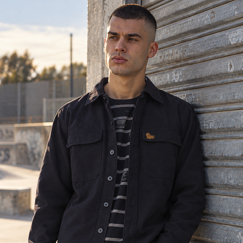 A man wearing a dark navy overshirt over a gray and black striped t-shirt, leaning against a corrugated metal shutter outdoors.