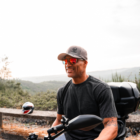 A man wearing a gray cap with a skull logo, red-tinted sunglasses, and a dark gray t-shirt sits on a motorcycle outdoors. A matching helmet rests on a wooden fence in the background against a hazy, overcast sky.