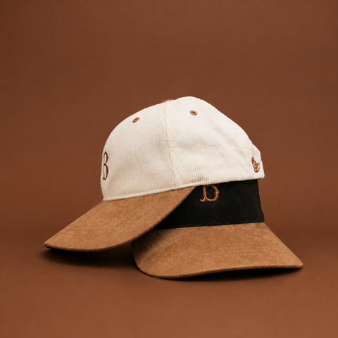 A structured baseball cap with an off-white corduroy crown, a brown felt visor, and a black fabric band featuring brown embroidered lettering, set against a solid brown background.