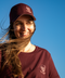 A smiling woman outdoors against a clear blue sky, wearing a burgundy baseball cap and matching burgundy T-shirt with a small white skull-and-cap logo and the text 'THE TRIPLET'.