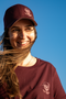 A smiling woman outdoors against a clear blue sky, wearing a burgundy baseball cap and matching burgundy T-shirt with a small white skull-and-cap logo and the text 'THE TRIPLET'.