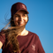 A smiling woman outdoors against a clear blue sky, wearing a burgundy baseball cap and matching burgundy T-shirt with a small white skull-and-cap logo and the text 'THE TRIPLET'.