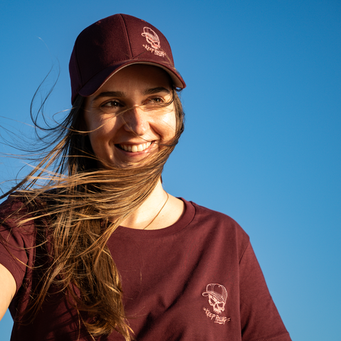 A smiling woman outdoors against a clear blue sky, wearing a burgundy baseball cap and matching burgundy T-shirt with a small white skull-and-cap logo and the text 'THE TRIPLET'.