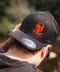 A man wearing a black baseball cap with an orange embroidered skull wearing a cap and Japanese katakana text, photographed outdoors with a blurred natural background.