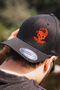 A man wearing a black baseball cap with an orange embroidered skull wearing a cap and Japanese katakana text, photographed outdoors with a blurred natural background.