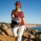A woman stands on rocky beach terrain holding a black motorcycle helmet, wearing a maroon t-shirt with a small white logo, a matching maroon cap, and white pants.