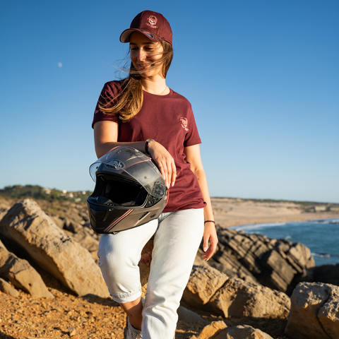 A woman stands on rocky beach terrain holding a black motorcycle helmet, wearing a maroon t-shirt with a small white logo, a matching maroon cap, and white pants.