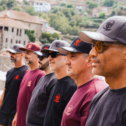 A group of men standing outdoors in a row, wearing matching black and burgundy t-shirts with a red skull logo and dark baseball caps, photographed in a scenic village setting.