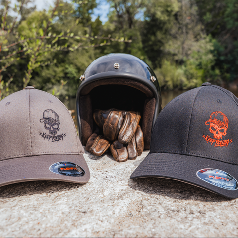 Two baseball caps displayed outdoors on a stone surface with a black motorcycle helmet and brown leather gloves behind them. The left cap is taupe with a black embroidered skull logo and text, and the right cap is black with an orange embroidered skull logo and text.
