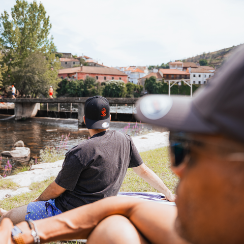 A man wearing a black baseball cap with a red logo and a dark gray t-shirt sits on a grassy bank by a river, with a bridge, water, and buildings in the background. Another person is blurred in the foreground.