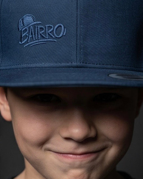 A close-up of a person wearing a dark navy blue snapback cap with an embroidered 'BAIRRO' logo on the front, set against a dark gray background.