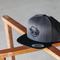 A dark gray heathered snapback cap with a black brim and a black embroidered skull wearing a hat on the front, resting on a wooden chair against a concrete floor.