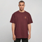 A man wearing an oversized maroon crewneck t-shirt with a small orange graphic on the left chest, photographed against a neutral light gray background.