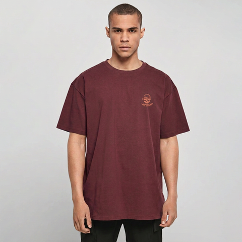 A man wearing an oversized maroon crewneck t-shirt with a small orange graphic on the left chest, photographed against a neutral light gray background.