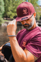 A man outdoors by a river wearing a maroon snapback cap and matching maroon t-shirt, both featuring a skull and crossbones logo with a helmet graphic. He is holding a black helmet in his other hand.
