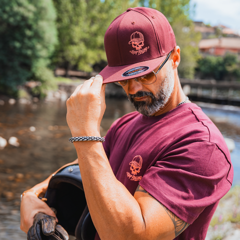 A man outdoors by a river wearing a maroon snapback cap and matching maroon t-shirt, both featuring a skull and crossbones logo with a helmet graphic. He is holding a black helmet in his other hand.