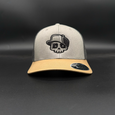 A structured trucker-style baseball cap with a light gray front panel, dark gray mesh back, and a tan visor. The front panel features an embroidered black and white skull wearing a cap.