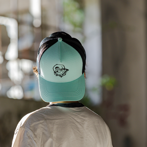 A person viewed from behind wearing a mint green and black trucker cap with a small black graphic logo on the front panel, in a softly blurred indoor setting.