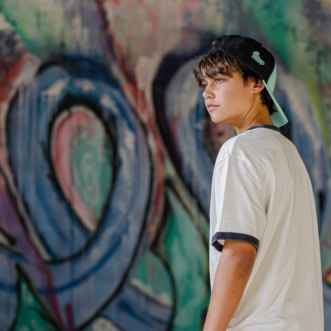 A young man wearing a white ringer t-shirt and a black and mint green baseball cap, standing in front of a colorful graffiti wall.