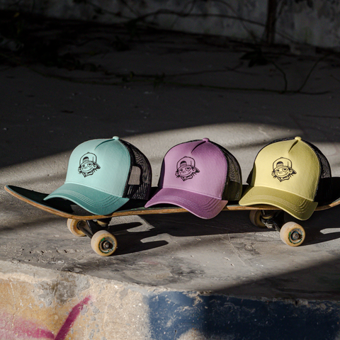 Three trucker-style baseball caps in mint green, lavender, and pale yellow are lined up on a skateboard outdoors on a concrete surface in shadowed sunlight.