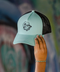 A mint green and black trucker hat with a black mesh back and a black embroidered cartoon graphic on the front, held by a hand against a blurred graffiti background.