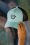 A mint green and black trucker hat with a black mesh back and a black embroidered cartoon graphic on the front, held by a hand against a blurred graffiti background.