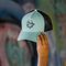 A mint green and black trucker hat with a black mesh back and a black embroidered cartoon graphic on the front, held by a hand against a blurred graffiti background.