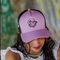 A person wearing a lavender trucker hat with black mesh side panels and a black cartoon-style graphic on the front. The person has dark curly hair, a white ruffled top, and a turquoise beaded necklace, photographed against a blurred outdoor background.
