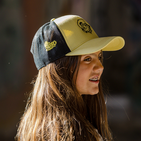 A woman in profile wearing a yellow and black trucker-style cap with embroidered logos, photographed outdoors against a blurred dark background.