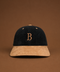 A structured baseball cap made of dark navy corduroy and tan suede, featuring an embroidered tan letter 'B' on the front. The cap is centered against a solid brown background.