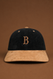 A structured baseball cap made of dark navy corduroy and tan suede, featuring an embroidered tan letter 'B' on the front. The cap is centered against a solid brown background.