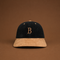 A structured baseball cap made of dark navy corduroy and tan suede, featuring an embroidered tan letter 'B' on the front. The cap is centered against a solid brown background.