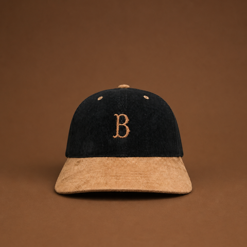 A structured baseball cap made of dark navy corduroy and tan suede, featuring an embroidered tan letter 'B' on the front. The cap is centered against a solid brown background.