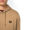 Close-up of a person wearing a tan hoodie with a small black embroidered graphic on the left chest, shown against a plain white background.