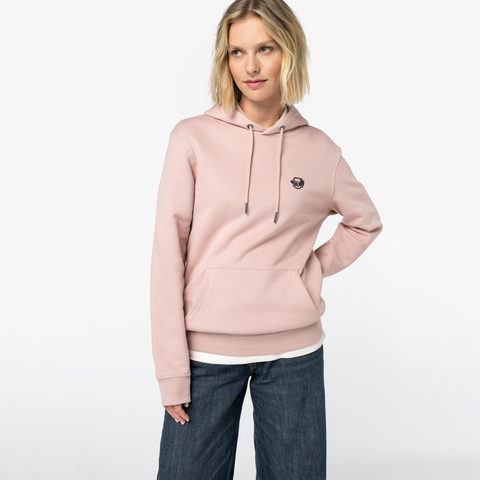 A woman modeling a light pink pullover hoodie with a small embroidered logo on the left chest, paired with dark blue jeans, against a plain white studio background.