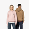 A man and a woman modeling pullover hoodies against a plain white studio background. The woman wears a light pink hoodie with a small black circular chest graphic, and the man wears a tan/camel hoodie from the back.