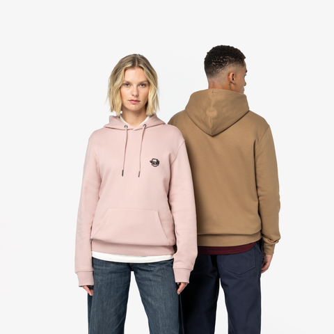 A man and a woman modeling pullover hoodies against a plain white studio background. The woman wears a light pink hoodie with a small black circular chest graphic, and the man wears a tan/camel hoodie from the back.