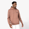 A man wearing a dusty rose pink pullover hoodie with a small black circular chest logo, paired with white pants, photographed against a clean white studio background.