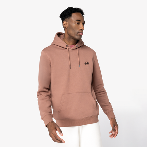 A man wearing a dusty rose pink pullover hoodie with a small black circular chest logo, paired with white pants, photographed against a clean white studio background.