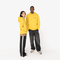 A man and a woman modeling matching bright yellow hoodies against a plain white studio background. The man wears dark gray jeans and beige sneakers, while the woman wears black track pants with a white side stripe and white sneakers.