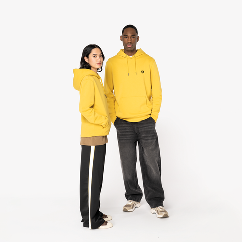 A man and a woman modeling matching bright yellow hoodies against a plain white studio background. The man wears dark gray jeans and beige sneakers, while the woman wears black track pants with a white side stripe and white sneakers.