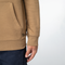 Close-up of a person wearing a tan hoodie with a kangaroo pocket and ribbed cuffs, paired with dark navy pants, against a light gray background.