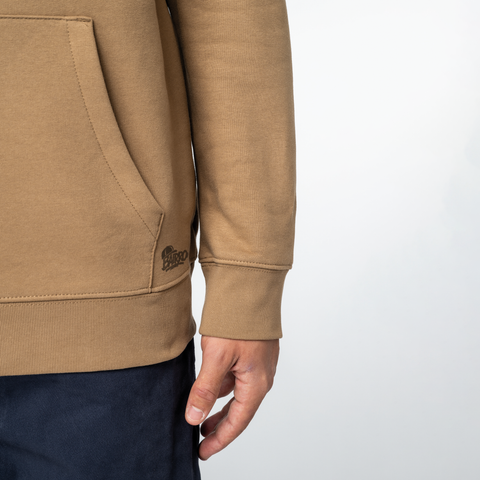 Close-up of a person wearing a tan hoodie with a kangaroo pocket and ribbed cuffs, paired with dark navy pants, against a light gray background.