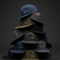 A stack of multiple baseball caps in navy, charcoal, olive, and black, each featuring a small white embroidered graphic on the front, set against a dark gray studio background.