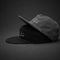 Two flat-brim caps stacked on a dark studio background. The top cap is charcoal gray and the bottom cap is black, both featuring a small white embroidered skull-and-crossbones style logo on the front.
