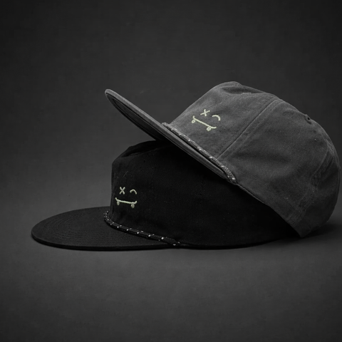 Two flat-brim caps stacked on a dark studio background. The top cap is charcoal gray and the bottom cap is black, both featuring a small white embroidered skull-and-crossbones style logo on the front.