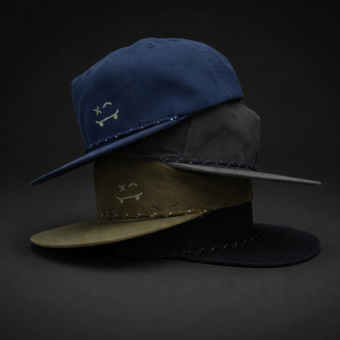 Three flat-brim cap hats stacked together on a dark studio background. The top hat is navy blue, the middle is olive green, and the bottom is black, each with a small embroidered skateboard face on the front and a row of metallic studs along the brim.