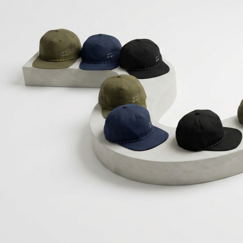 Six baseball caps in olive green, navy blue, and black, arranged on a sculptural white display stand against a clean white studio background.