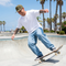 A man wearing a white t-shirt, light blue jeans, black sneakers, and an olive green cap is skateboarding in a concrete bowl at a beach skatepark with palm trees in the background.