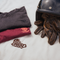 Two folded graphic T-shirts, one dark gray and one burgundy, displayed on a white fabric surface next to a black motorcycle helmet and brown leather gloves. The burgundy shirt features a skull graphic with the text 'Krazy Speedway' and the word 'BAIRRO' is embroidered on the white fabric below.